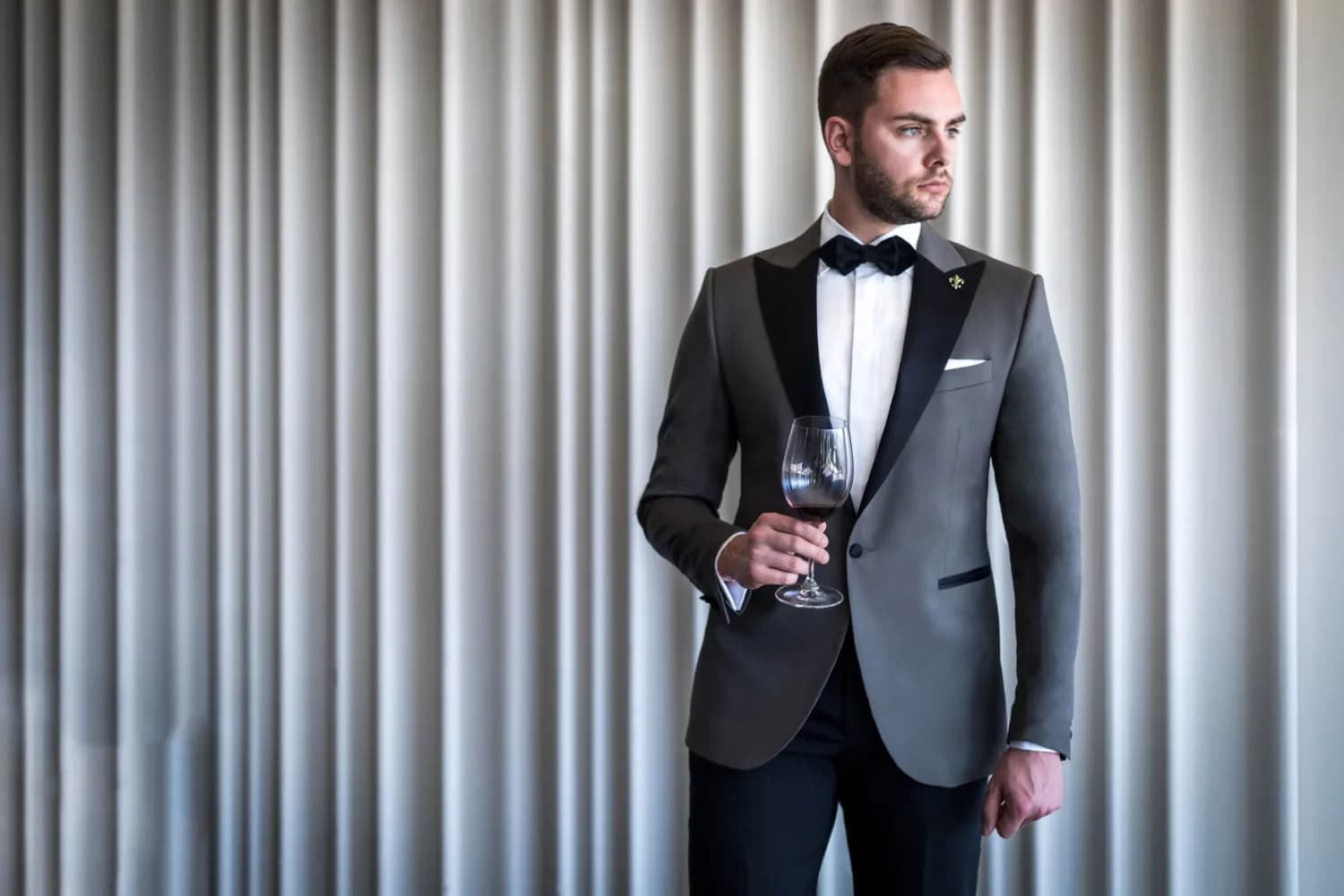 Quý ông mặc tuxedo xám ve satin đen cầm ly rượu trước rèm sáng