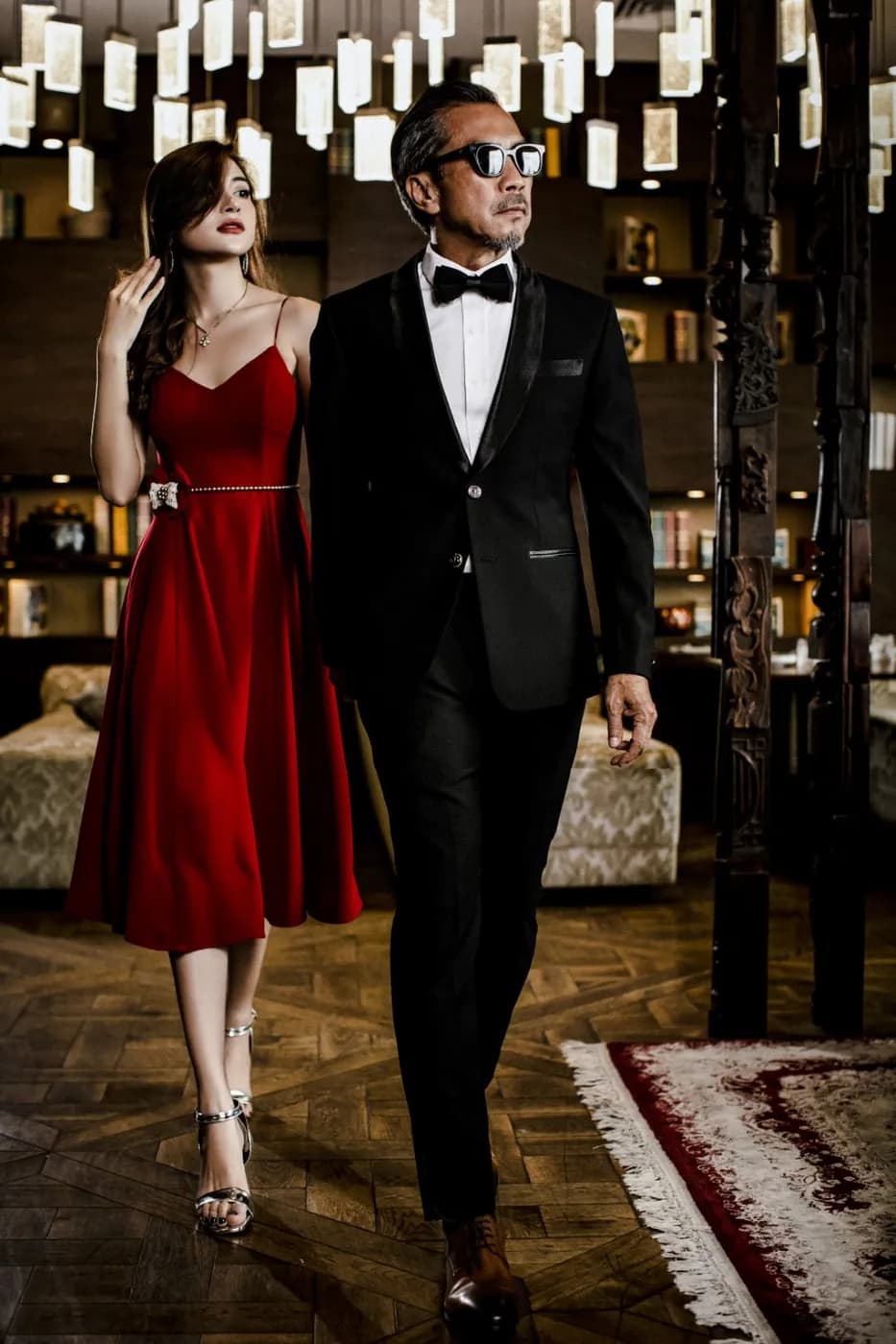 Quý ông mặc tuxedo đen sánh bước cùng người phụ nữ váy đỏ trong sảnh tiệc